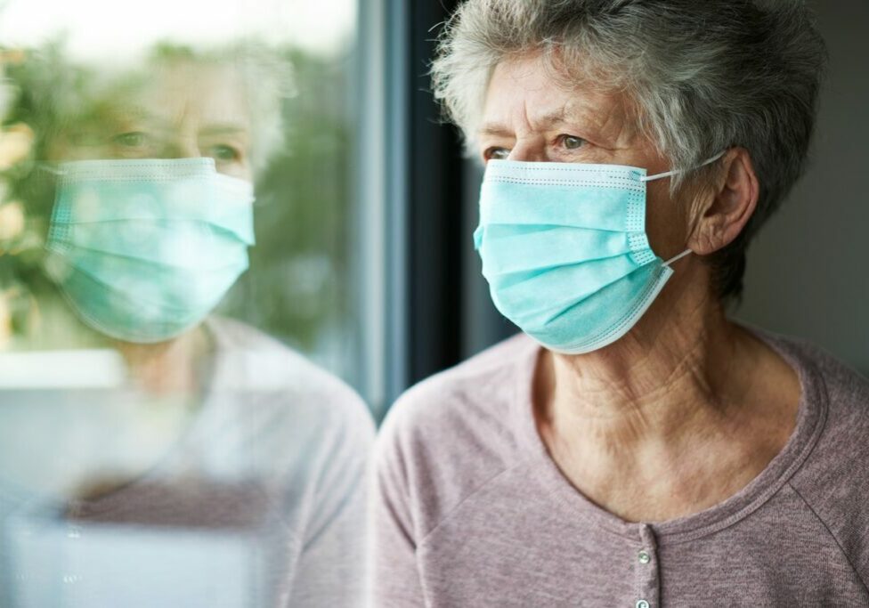 a old woman or grandma is wearing a respirator or surgical mask and looking out of the window inactive and isolated