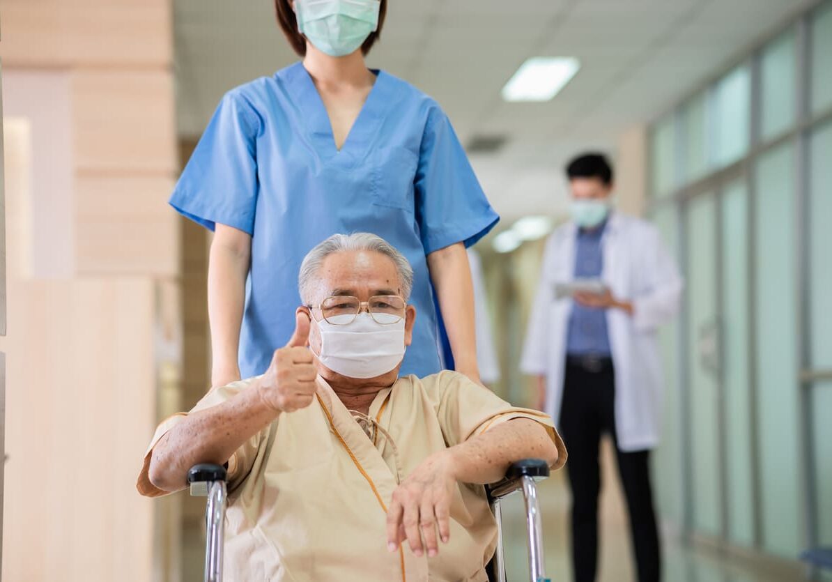 Portrait of senior Asian man wear protective face mask sitting on wheelchair and recovering from illness showing thumbs up and looking at camera while nurse caring taking him for relax