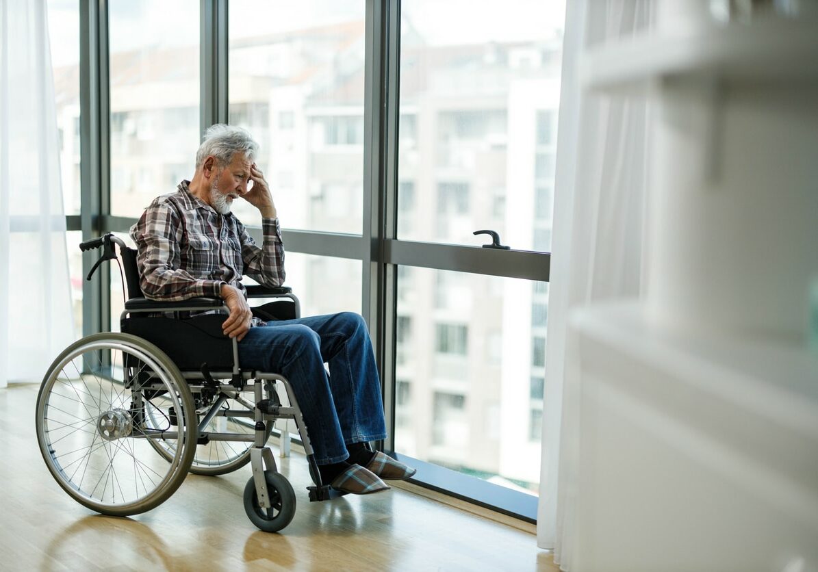 Sad senior man in a wheelchair looking through window from nursing home.