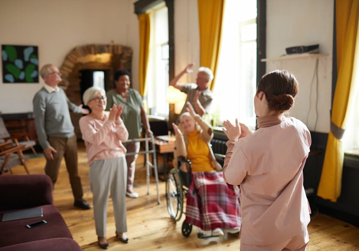 Diverse group of smiling senior people dancing while enjoying activities in retirement home with young woman in foreground, copy space