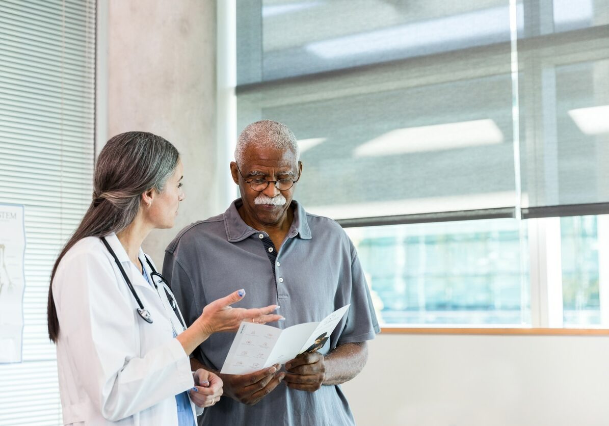 A mid adult female doctor gestures as she discusses home healthcare options with a senior adult male patient. The man is reading a home healthcare informational brochure.