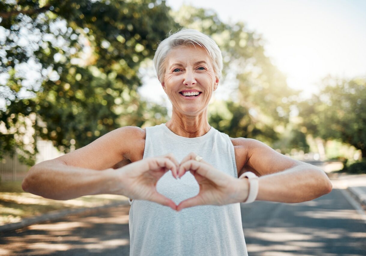 Fitness, happy and heart hands of old woman in nature after running for health, wellness and workout. Smile, motivation and peace with senior lady and sign for love, faith and training in nature